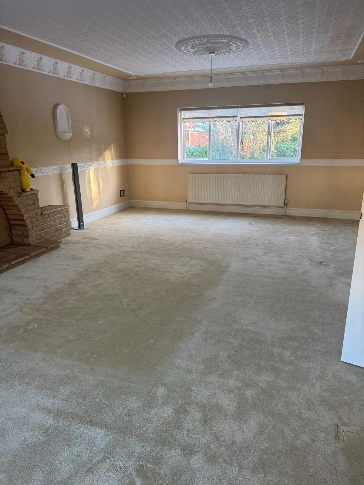 Empty room under renovation with beige walls, concrete floor, fireplace, radiator, and large window with autumn view