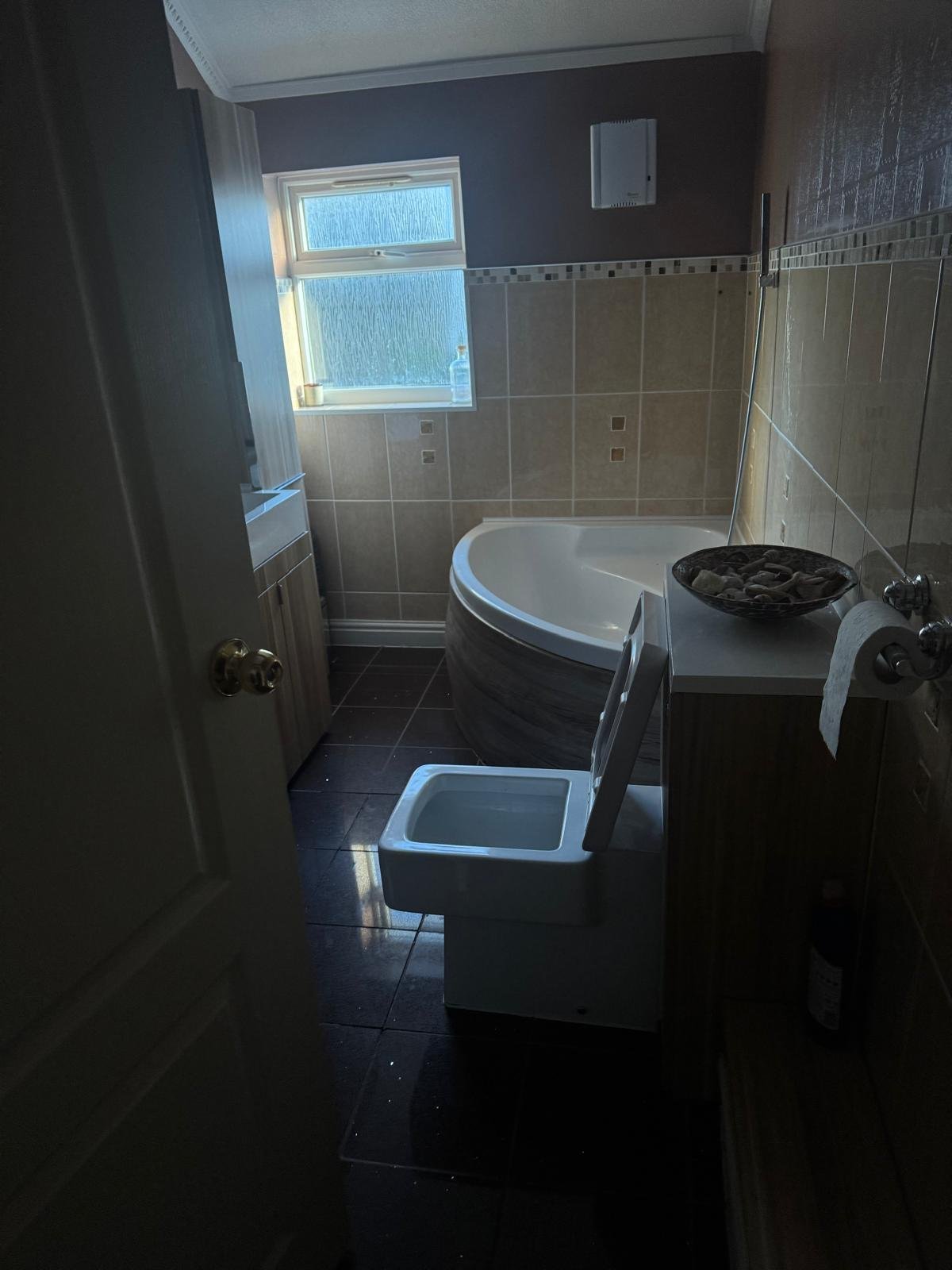 Bathroom with beige and dark tile walls, white bathtub, blue bath mat, window, and dark flooring