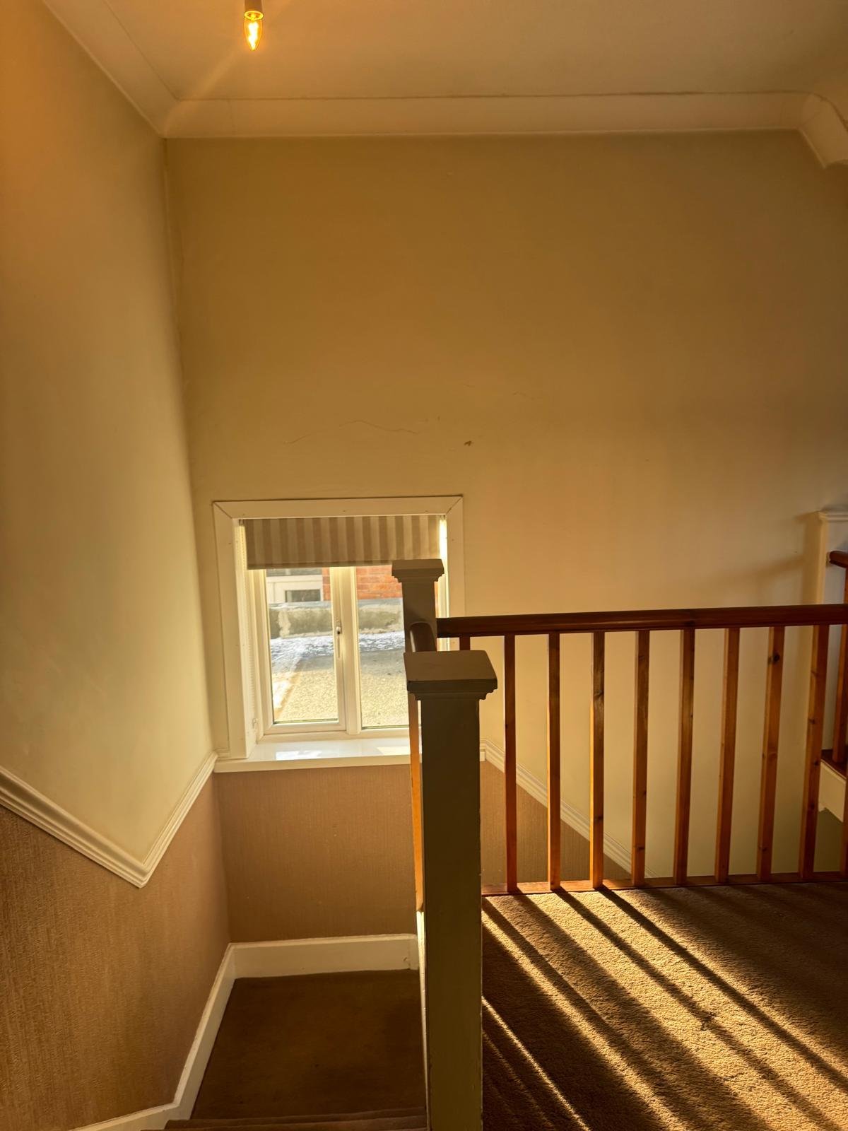 Sunlit staircase landing with wooden railing, cream walls, and window letting in natural light