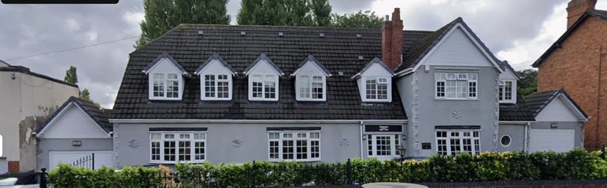 Two-story gray and white house with mansard roof, dormer windows, brick chimneys, and manicured hedge in front